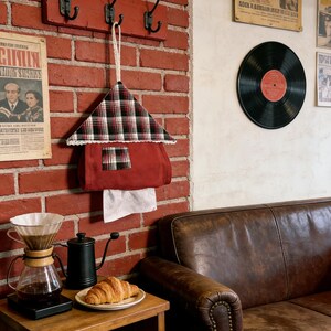 May include: A red brick wall with a wooden coat rack featuring three black hooks. A plaid fabric house-shaped tissue holder hangs below. A coffee setup and a leather couch are also visible.