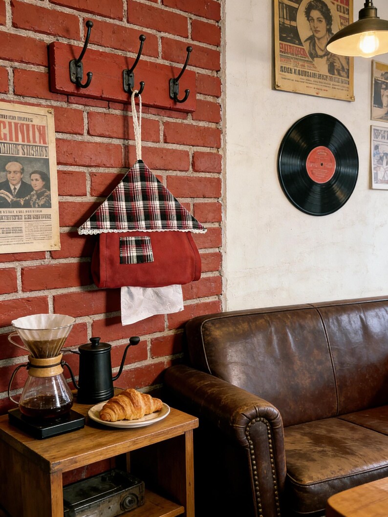 May include: A red brick wall with a wooden coat rack featuring three black hooks. A plaid fabric house-shaped tissue holder hangs below. A coffee setup and a leather couch are also visible.