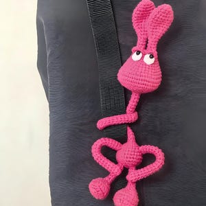 May include: A bright pink, crocheted bunny toy with large, wide eyes and long, floppy ears. The bunny has a long, flexible body and limbs, and is attached to a dark gray surface. The toy is handmade and has a whimsical design.