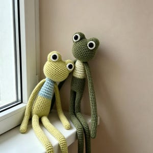 May include: Two handmade crochet frog dolls with long limbs and large eyes. The left frog is light yellow with a light blue chest patch. The right frog is dark green with a light green chest patch. Both have round heads and long, dangling legs.