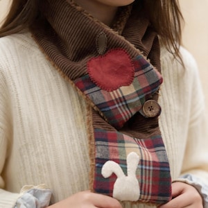 May include: Brown corduroy scarf with a plaid pattern, a red apple patch, and a white bunny applique. The scarf has a button closure and a soft, fuzzy lining. A cozy accessory for fall and winter.