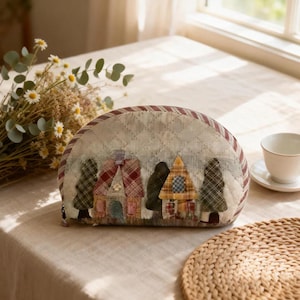 Könnte beinhalten: Eine gesteppte, halbmondförmige Tasche mit einer Dorflandschaft aus Häusern und Bäumen. Die Tasche besteht aus gemusterten Stoffen in Rot-, Grün- und Beigetönen, mit einem Steppmuster und einem Reißverschluss.