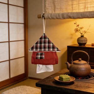 May include: A red and plaid fabric tissue box cover shaped like a house, hanging from a string. The house-shaped cover has a plaid roof and a small window. White tissues are visible at the bottom. The scene is set in a traditional Japanese-style room.
