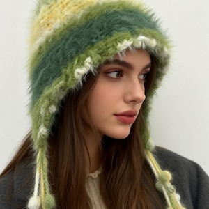 May include: A fuzzy, colorful hat with ear flaps and dangling tassels. The hat features a gradient of yellow, green, and white. The tassels are adorned with small, fluffy pom-poms. The hat is made of a soft, textured material, perfect for cold weather.