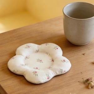 May include: A white, flower-shaped coaster with a floral pattern of small pink roses and green leaves. A beige ceramic mug sits next to the coaster on a wooden surface. The coaster is a decorative item.