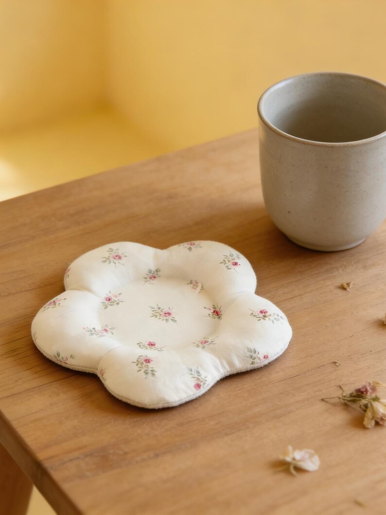 May include: A white, flower-shaped coaster with a floral pattern of small pink roses and green leaves. A beige ceramic mug sits next to the coaster on a wooden surface. The coaster is a decorative item.
