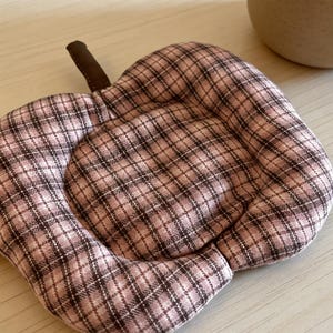 May include: A quilted, apple-shaped trivet with a brown stem. The trivet features a pink and brown plaid pattern. The trivet is designed to protect surfaces from hot dishes and is a decorative kitchen accessory.
