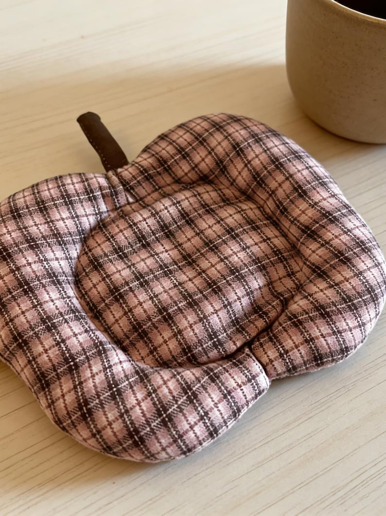 May include: A quilted, apple-shaped trivet with a brown stem. The trivet features a pink and brown plaid pattern. The trivet is designed to protect surfaces from hot dishes and is a decorative kitchen accessory.