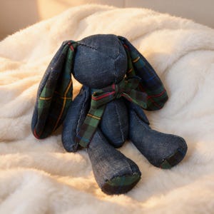 May include: A denim bunny plush toy with plaid detailing on the ears and bow. The bunny is a dark blue colour, with a green, red, and yellow plaid pattern. The toy is resting on a soft, white, fluffy surface.