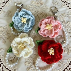 May include: Four crochet flower keychains in blue, pink, white, and red. Each flower has a yellow center, green leaf, and a pearl bead wristlet. The keychains have silver-colored clasps.