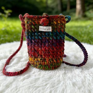 May include: A handmade, rainbow-colored crochet bag with a long, braided strap. The bag features a button closure and a small tag that reads "Handmade". The bag is sitting on a white, fluffy surface.