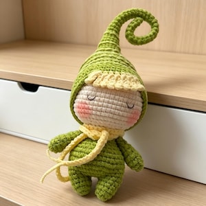 May include: A small, handmade crochet doll featuring a green pointed hat with a curled tip, a pale beige face with closed eyes and pink cheeks, and a green body. The doll wears a light yellow brim under the hat and a matching yellow scarf.