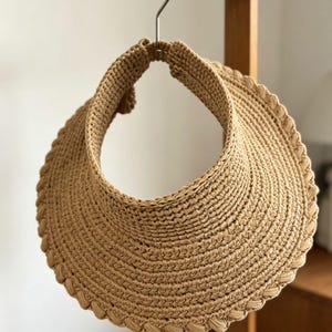 May include: A tan crochet sun visor featuring a wide, circular brim with a braided edge detail. The visor is constructed from a textured, woven material and includes an open top design, hanging from a metal hook on a wooden rod.