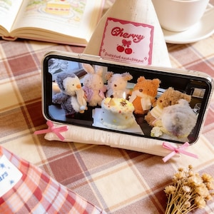 May include: A phone displaying a photo of plush toys around a cake, resting on a white pillow with pink ribbons. A book, a cup of coffee, and a fabric triangle with "Cherry" text are also visible on a plaid surface.