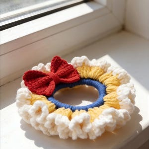 May include: A handmade crochet hair scrunchie featuring a red bow, yellow and white ruffled trim, and a blue center. This unique hair accessory adds a touch of handmade charm to any hairstyle.