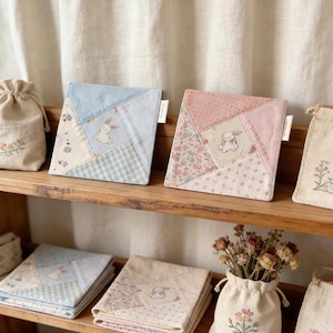 Puede incluir: Posavasos cuadrados de tela acolchada con dise&ntilde;os de patchwork. Cada posavasos presenta un patr&oacute;n diferente, incluyendo estampados florales, cuadros de vichy y colores lisos. Un peque&ntilde;o conejo bordado est&aacute; centrado en cada posavasos. Los posavasos se exhiben en una estanter&iacute;a de madera.