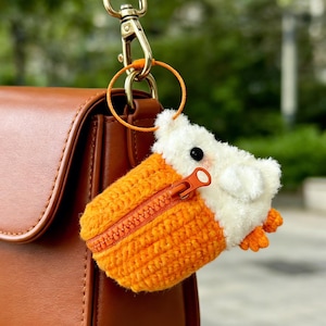 May include: A handmade orange and white crochet keychain in the shape of a small animal, with a zipper closure. The keychain is attached to a brown leather purse with a gold-colored clasp.