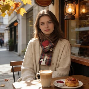 May include: A woman wearing a cream cardigan and a plaid scarf with a bunny design sits at a cafe table. A mug of coffee, a plate with a pastry, and a small dish of jam are on the table. Autumn leaves are scattered around.