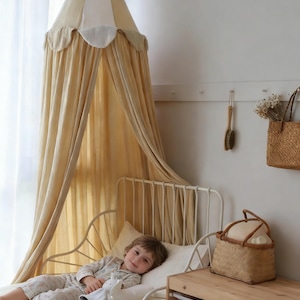 May include: A child's bedroom scene featuring a cream-colored canopy bed with a metal frame. The bed is adorned with a soft, light-colored bedding set. A woven basket and a small wooden storage unit are nearby. The room has a neutral color palette.