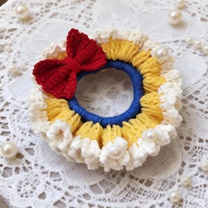 May include: A handmade crochet hair tie featuring a red bow, yellow and white trim, and a blue center. The hair accessory is adorned with faux pearls, creating a whimsical and decorative design.