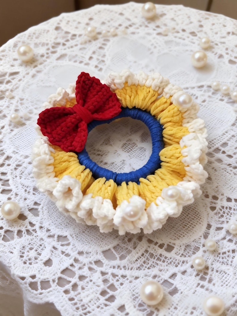 May include: A handmade crochet hair tie featuring a red bow, yellow and white trim, and a blue center. The hair accessory is adorned with faux pearls, creating a whimsical and decorative design.