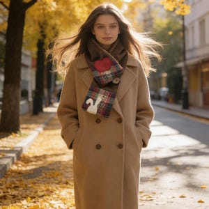 May include: A woman in a tan double-breasted coat and a scarf with a red heart and plaid pattern. The scarf is brown and the coat is knee-length. The background shows a street with yellow leaves and trees.
