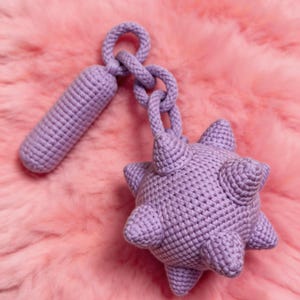 May include: A lavender crocheted toy mace with a cylindrical handle, a chain, and a spiky ball head. The toy is made of soft yarn and is designed for play. The background is a pink, fluffy surface.
