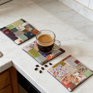 May include: Three handmade patchwork coasters in various colors and patterns, with a clear glass cup of coffee on one. Coffee beans are scattered nearby. The coasters are square and appear to be made of fabric scraps.