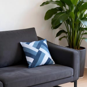 May include: A gray sofa with a blue and white geometric patterned pillow. A large potted plant with green leaves is in the background. The sofa has a modern design with black metal legs.