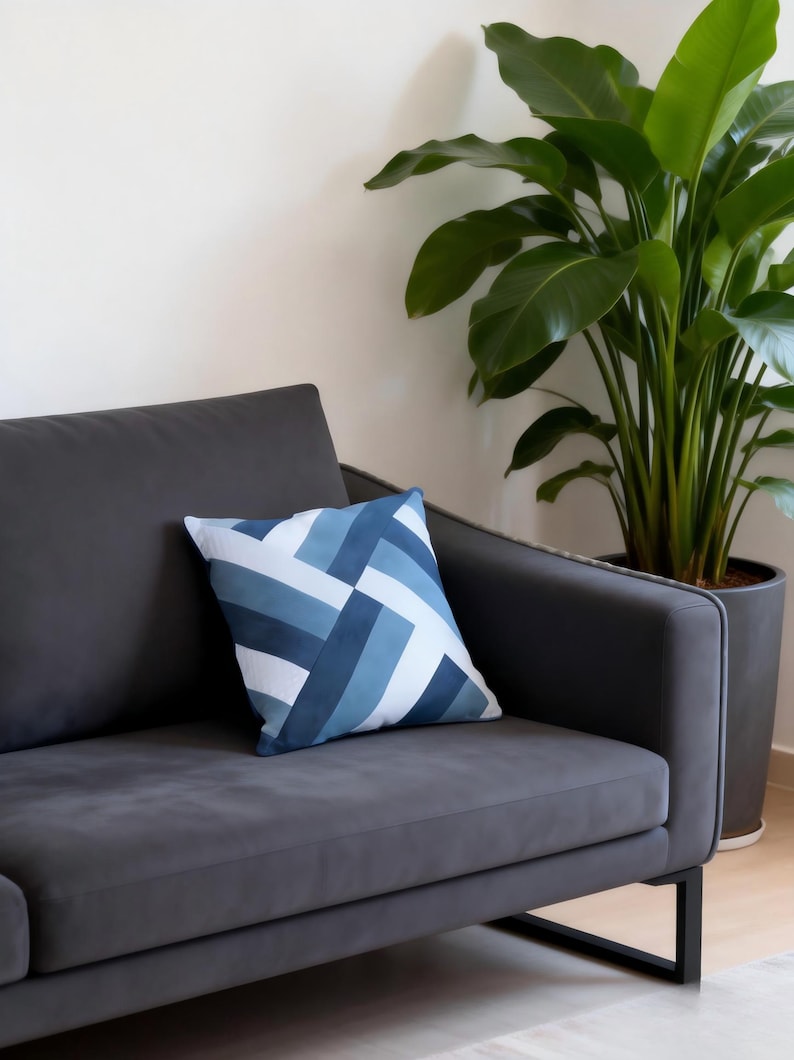 Op de afbeelding: Een grijze bank met een blauw en wit geometrisch patroon kussen. Op de achtergrond staat een grote potplant met groene bladeren. De bank heeft een modern design met zwarte metalen poten.