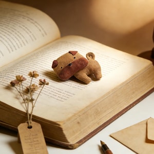 May include: A plush pug dog toy in shades of brown and tan rests on an open book. The toy has a detailed face and a curled tail. A small bouquet of dried flowers with a tag and a pen are also on the table.
