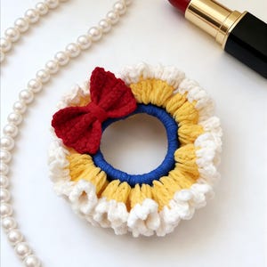 May include: A handmade crochet hair scrunchie with a red bow, yellow, white, and blue accents. A pearl necklace and red lipstick are also in the image. This hair accessory is perfect for adding a touch of handmade charm to any hairstyle.
