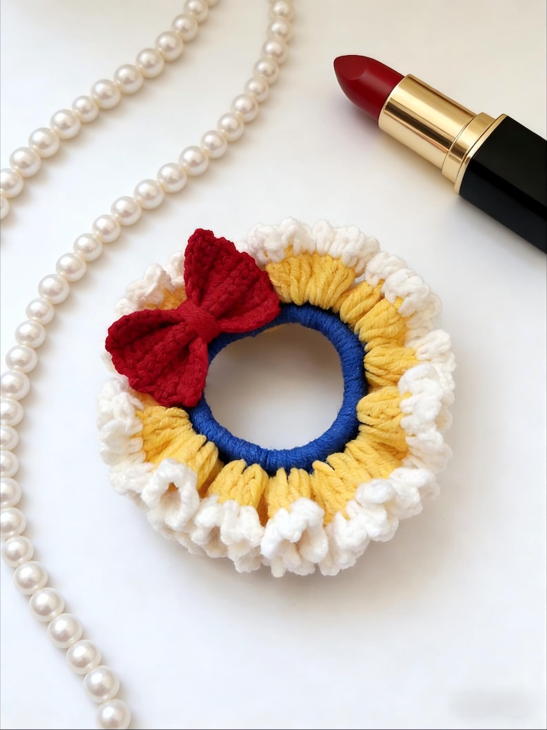 May include: A handmade crochet hair scrunchie with a red bow, yellow, white, and blue accents. A pearl necklace and red lipstick are also in the image. This hair accessory is perfect for adding a touch of handmade charm to any hairstyle.