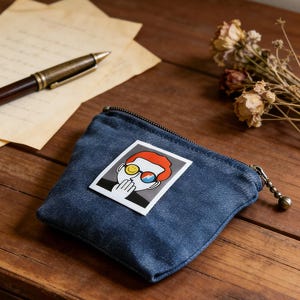 Puede incluir: Un monedero de mezclilla con cierre de cremallera y una pequeña bola de metal adjunta. El monedero presenta un gráfico colorido de una persona con gafas de sol. El monedero está sobre una superficie de madera con un bolígrafo y flores secas.