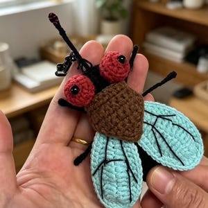 Puede incluir: Un llavero de mosca tejido a ganchillo con cuerpo marrón, dos grandes ojos rojos con pupilas negras y alas azul claro con detalles de venas bordadas en negro. El insecto tiene antenas y patas negras finas, con una anilla de metal negro arriba.