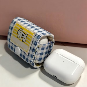 May include: A white AirPod case next to a blue and white gingham AirPod case cover. The cover has a yellow patch with a cartoon dog illustration. The AirPod case is open, revealing the white earbuds.