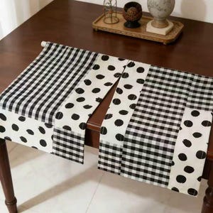 May include: A black and white patterned table runner. The runner features alternating sections of gingham check and polka dots. The runner is displayed on a dark brown wooden table.