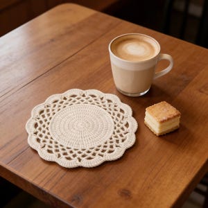 Puede incluir: Un posavasos de ganchillo color crema sobre una mesa de madera, junto a una taza de café beige y un pequeño pastel cuadrado. El posavasos tiene un centro circular y un borde festoneado. El café tiene un diseño en forma de corazón.