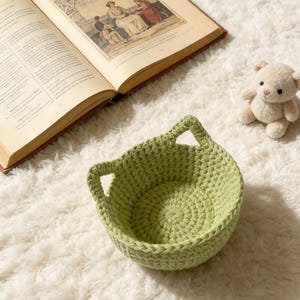 May include: A light green, handmade crochet basket with ear-shaped handles sits on a fluffy white rug. An open book with illustrations and a small teddy bear are also in the image. The basket is round and has a textured appearance.