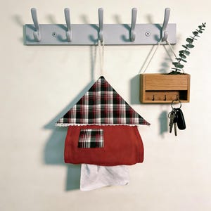 May include: A wall-mounted organizer featuring a gray coat rack with hooks, a wooden key holder, and a decorative house-shaped tissue box. The tissue box is red with a plaid roof and a white lace trim.