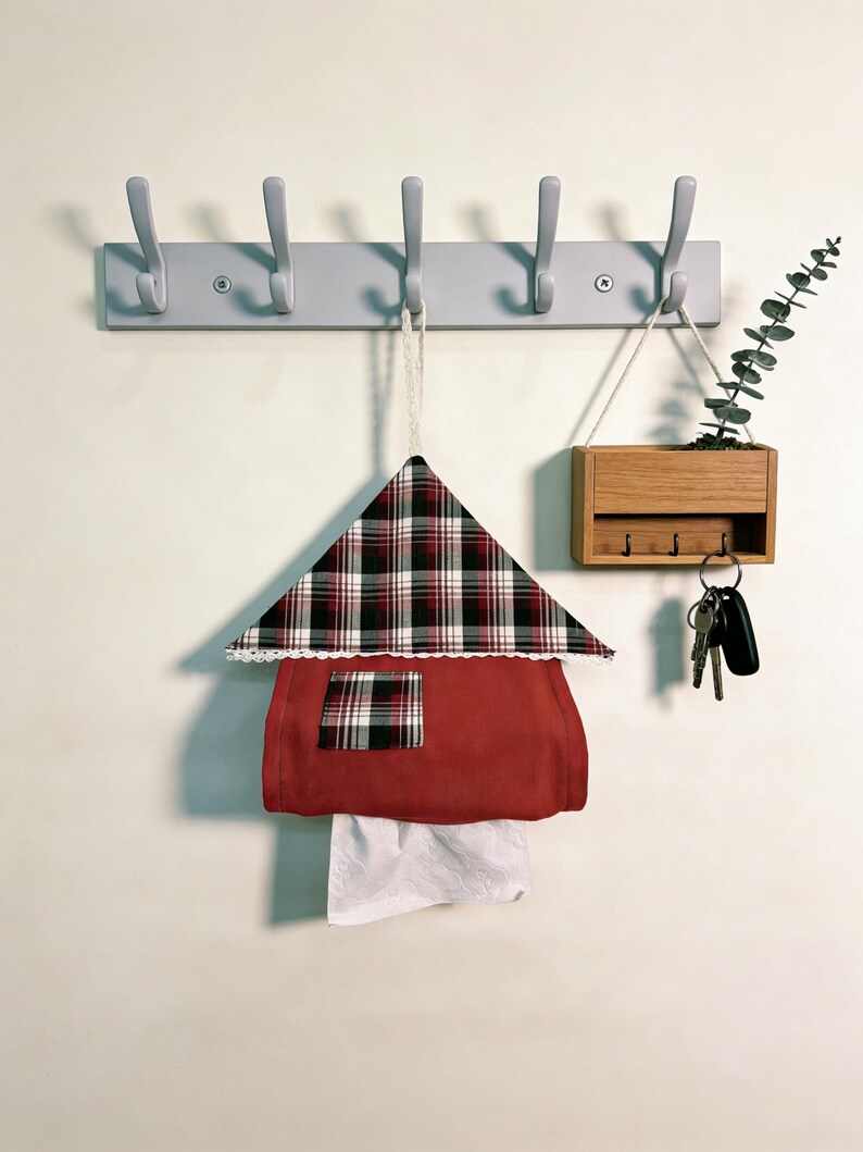 May include: A wall-mounted organizer featuring a gray coat rack with hooks, a wooden key holder, and a decorative house-shaped tissue box. The tissue box is red with a plaid roof and a white lace trim.