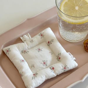 以下が含まれることがあります： 小さな赤と緑のバラのフローラルパターンが施された、白の正方形のキルティングコースター。コースターは、レモンスライスが入った水のグラスの隣のピンクのトレイに置かれています。