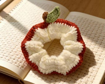 May include: A handmade crochet scrunchie in the shape of an apple. The scrunchie features a red exterior, a white center, and a green leaf and brown stem detail. The scrunchie is resting on an open notebook.