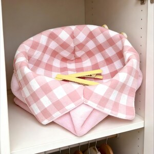Può includere: Un cestino in tessuto rosa e bianco a quadretti con due mollette gialle all'interno, appoggiato su un ripiano bianco. Il cestino è all'interno di un armadio bianco con un'asta di legno e grucce.
