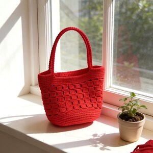 Op de afbeelding: Een felrode gehaakte tote bag met een geweven patroon staat op een vensterbank. De tas heeft een stevige handgreep en een gestructureerde vorm. Een kleine plant in een pot staat naast de tas en een bol rood garen is zichtbaar.