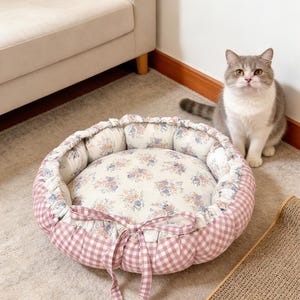 Könnte beinhalten: Ein rundes Katzenbett mit einem Kissen mit Blumenmuster und einer rosa Vichy-Außenseite. Das Bett hat einen gerüschten Rand und ist mit einer rosa Schleife gebunden. Eine grau-weiße Katze sitzt in der Nähe.