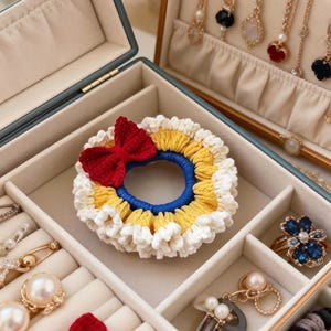 May include: A handmade crochet hair scrunchie with a red bow, yellow and white trim, and a blue center. The scrunchie is displayed in a jewelry box with various earrings, rings, and other accessories.