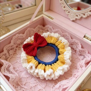 May include: A handmade crochet scrunchie with a red bow, yellow and white trim, and a blue center. The scrunchie is displayed inside a pink jewelry box with a lace lining and a decorative mirror.