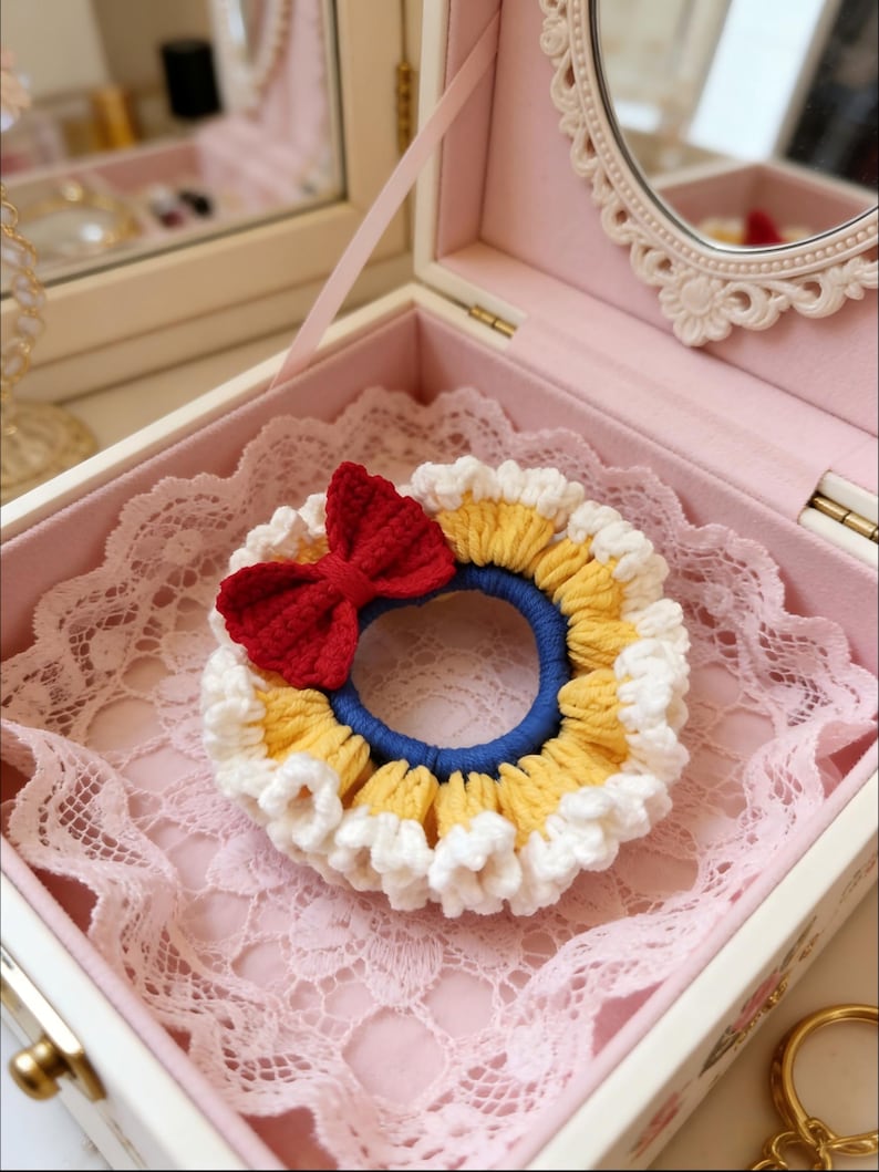 May include: A handmade crochet scrunchie with a red bow, yellow and white trim, and a blue center. The scrunchie is displayed inside a pink jewelry box with a lace lining and a decorative mirror.