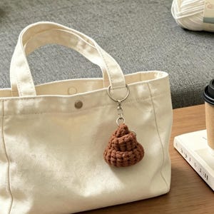 May include: A cream-colored canvas tote bag with two top handles and a metal snap closure. A brown crocheted keychain in a rounded, textured shape is attached to one of the bag's handles by a silver-toned metal ring and chain link hardware.
